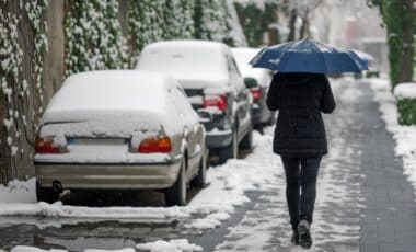 une personne vetue en noir qui porte un parapluie et marche dans une route enneigée, devant des voitures couvertes de neige