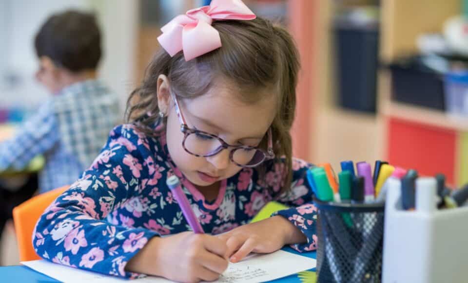 une petite fille qui porte des lunettes et un noeud papillon dans les cheveux, en train de dessiner dans ce qui semble etre une salle de classe pour évoquer l'allocation de rentrée scolaire