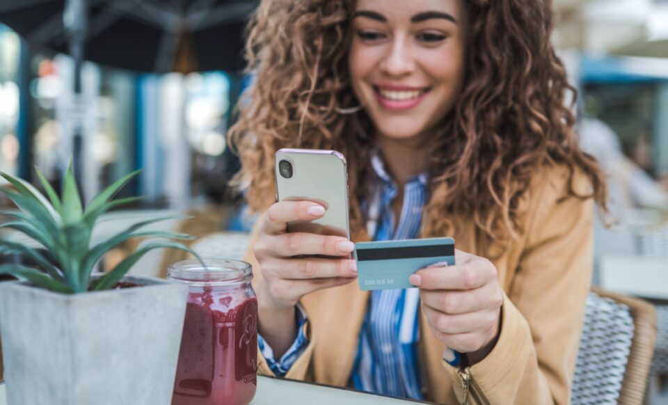 une jeune femme au cheveux bouclés tenant une carte bancaire dans une main et un smartphone dans l'autre main, assise dans ce qui semble etre la terasse d'un café