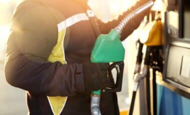 L'image montre un individu tenant un pistolet de pompe à essence dans une station-service, en train de faire le plein de carburant.