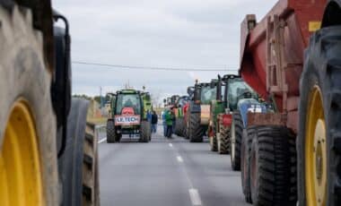 Cette image montre une file de tracteurs alignés sur une route, semblant participer à une mobilisation ou un blocage. Des personnes se tiennent entre les véhicules, ce qui évoque une action collective initiée par des agriculteurs.