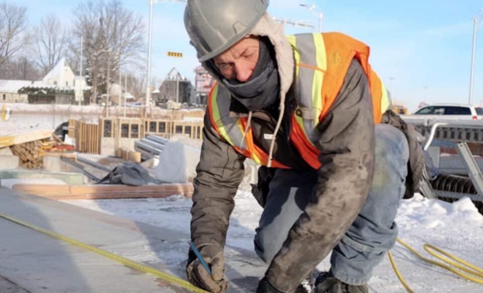 Un ouvrier du bâtiment travaille à l'extérieur dans des conditions hivernales, vêtu d'équipements adaptés au froid, sur un chantier enneigé.