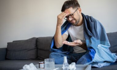 Un homme, enroulé dans une couverture, semble malade et prend des médicaments. L'image illustre les symptômes de la grippe et la prise en charge à domicile.