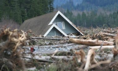 Une maison partiellement submergée et entourée de débris de bois, probablement à la suite d'une inondation ou d'une coulée de boue, dans un paysage forestier.