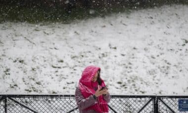 Une femme emmitouflée dans une écharpe rose consulte son téléphone sous la neige, avec un paysage enneigé en arrière-plan. Mot clé : météo.