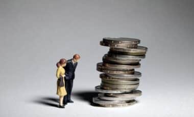 Deux figurines miniatures représentant un couple observent une pile instable de pièces de monnaie, illustrant l’incertitude économique et le moral des ménages face aux finances.