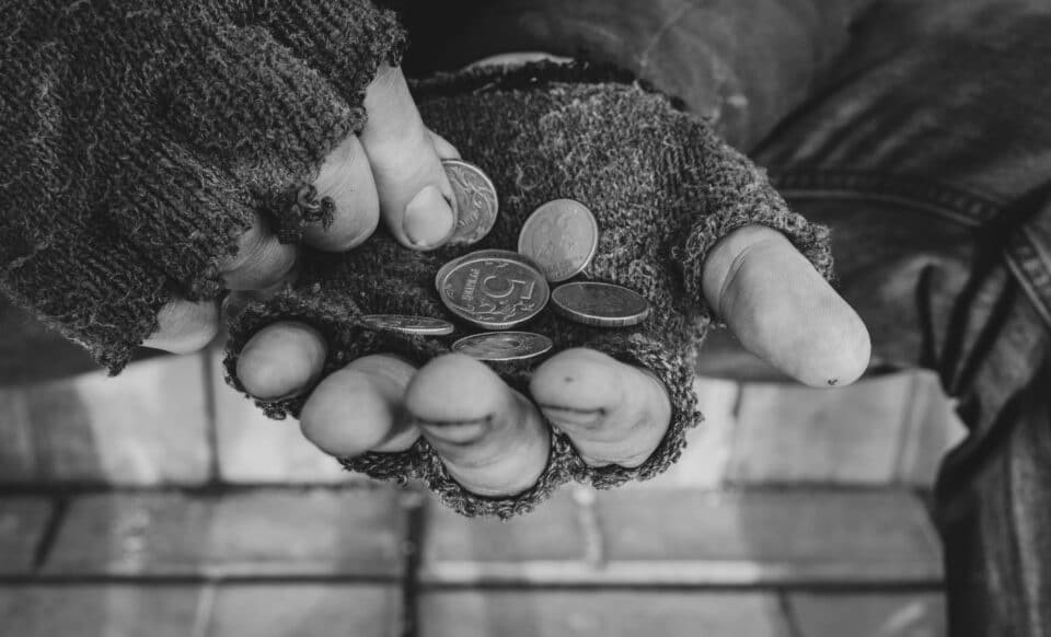 L'image montre une personne portant des gants usés, tenant quelques pièces de monnaie, symbolisant la pauvreté et la précarité financière.