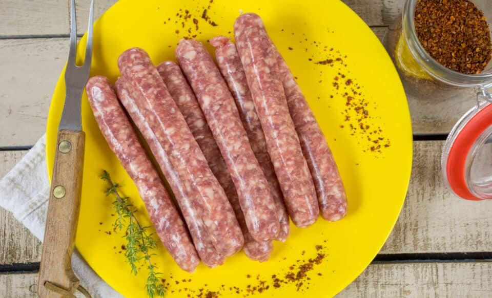 L'image montre des saucisses crues disposées sur une assiette jaune, accompagnées d'herbes et d'épices, prêtes pour la préparation culinaire.