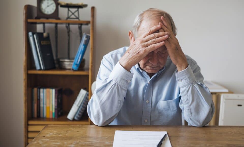 Cette image illustre un retraité visiblement préoccupé, assis à une table avec des documents devant lui, symbolisant des préoccupations liées à la retraite et à sa gestion financière.