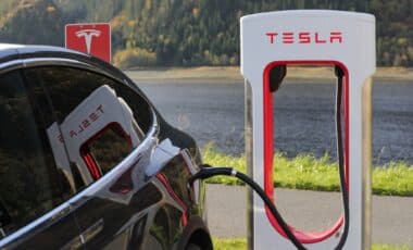 Cette image montre une voiture Tesla branchée à une borne de recharge Tesla, avec un cadre naturel en arrière-plan, comprenant des arbres et un plan d'eau.