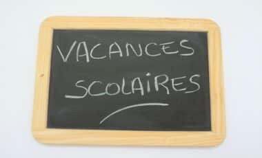 L'image montre une ardoise en bois sur laquelle est inscrit "Vacances scolaires" à la craie blanche, évoquant le thème des congés scolaires.