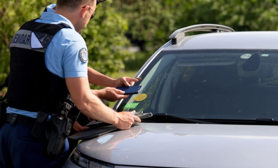 L'image montre un contrôle routier effectué par un gendarme vérifiant les documents d'un véhicule. Sur le pare-brise, une vignette Crit'Air est visible. Cette scène illustre l'importance des vignettes apposées sur les véhicules, notamment pour circuler dans certaines zones réglementées.