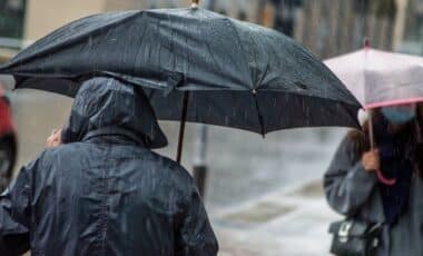 une personne dans la rue, de dos, vetue d'un impermeable noir, qui marche sous la pluie, tenant un parapluie noir