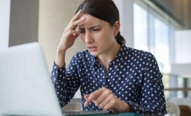 Une femme, cheveux noirs et chemise bleue à petits pois blancs, consulte son ordinateur, l'air inquiet pour illustrer les arnaques sur le Net