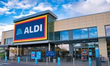 L'image montre l'entrée d'un magasin ALDI moderne, avec un logo bien visible sur la façade et un ciel bleu en arrière-plan.