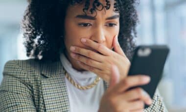 Une femme regarde son téléphone avec une expression inquiète, la main sur la bouche, suggérant qu'elle vient de découvrir une arnaque ou une mauvaise nouvelle en ligne.