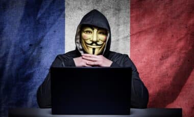 Un individu masqué, portant une capuche, est assis devant un ordinateur sur fond de drapeau français, symbolisant les cyberattaques et le piratage informatique visant la France.