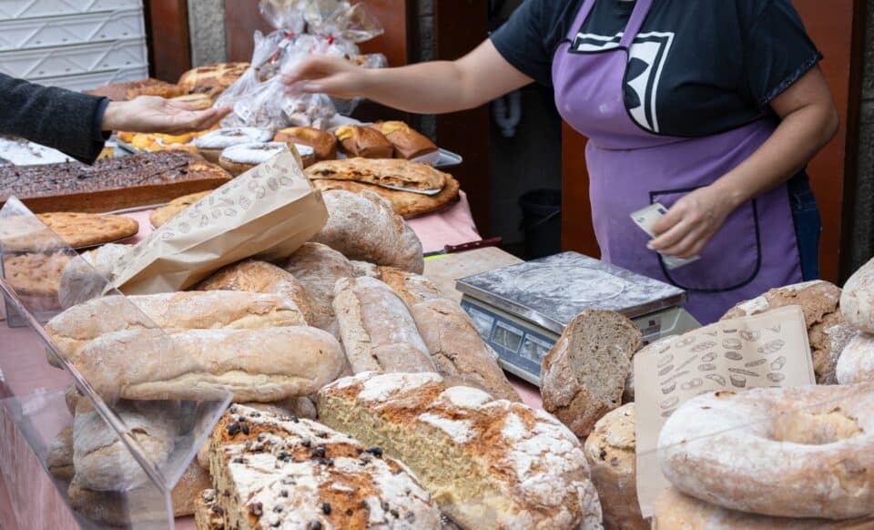Une boulangère sert un client sur un stand rempli de pains et pâtisseries, illustrant le quotidien des boulangers face aux nouvelles contraintes.