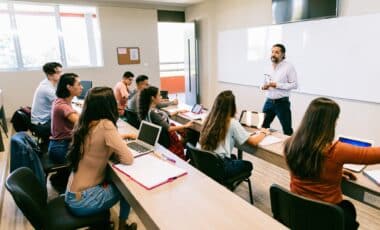 Un professeur donne un cours à un groupe d'étudiants attentifs, équipés d'ordinateurs et de carnets dans une salle de classe moderne.