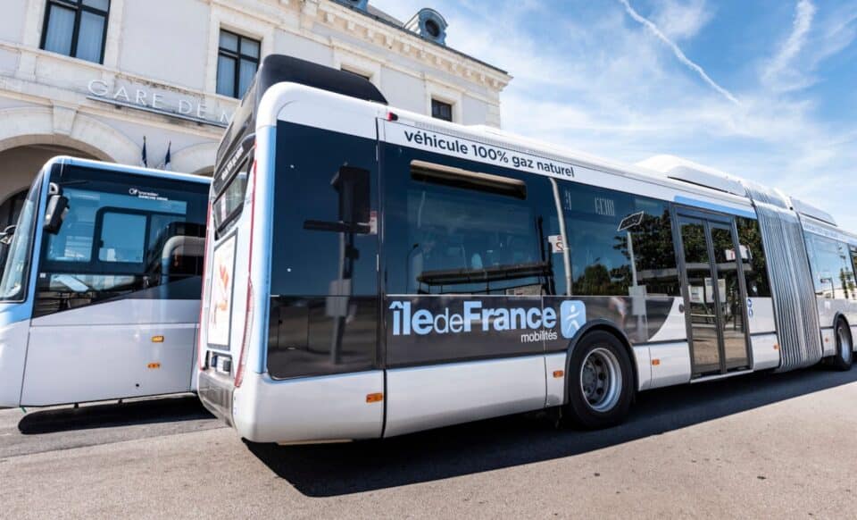 L'image montre un bus articulé Île-de-France Mobilités stationné devant une gare. Un autre bus est visible en arrière-plan. Le ciel est dégagé et ensoleillé.