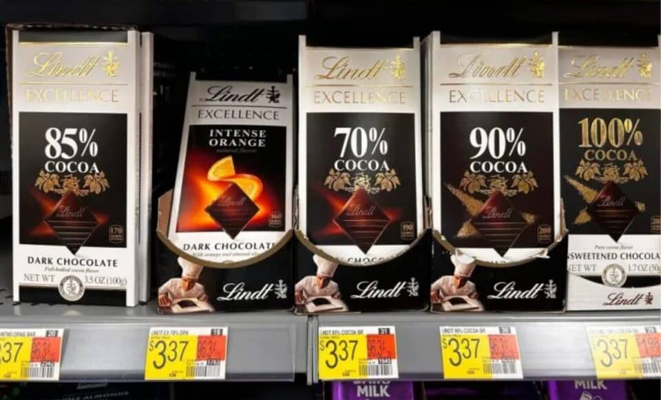 L'image montre des tablettes de chocolat Lindt Excellence en différentes variétés de cacao (70%, 85%, 90%, 100%) et saveur (intense orange), avec des prix affichés en magasin.