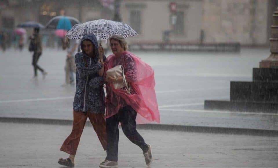 Deux femmes marchent sous la pluie, protégées par des parapluies, dans une scène illustrant des conditions météorologiques pluvieuses. Météo maussade et grise.