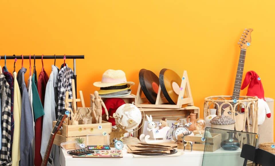 L'image montre un vide-grenier avec divers objets à vendre, tels que des vêtements, des vinyles, des instruments de musique, des jouets, des accessoires et des objets décoratifs, tous étiquetés avec des prix.